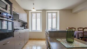 A kitchen with a built-in oven, microwave, and coffee maker. There are two windows with blinds, a hanging light fixture, and a table with chairs. - La Maison d'à côté - CRANCEY (CRANCEY)