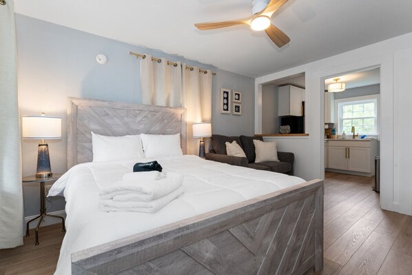 A hotel room with a large bed, a wooden headboard, a ceiling fan, a nightstand with a lamp, a seating area with a sofa, and a view of the kitchen.
