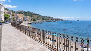 Une promenade en bord de mer, avec un chemin pavé de pierres, une rambarde en métal et une vue sur la mer, où l’on peut apercevoir des bateaux.