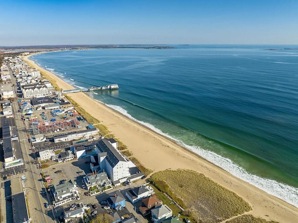 An aerial view of a coastal town with a beach, a pier, and a row of buildings along the shore.