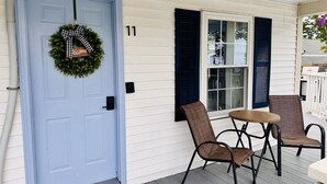 Eine Veranda mit einer blauen Tür, zwei Korbstühlen, einem kleinen runden Tisch und einem Willkommen-Kranz.