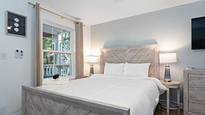 A bedroom with a wooden headboard, a large bed, two bedside tables with lamps, a small dresser, and a window with curtains.