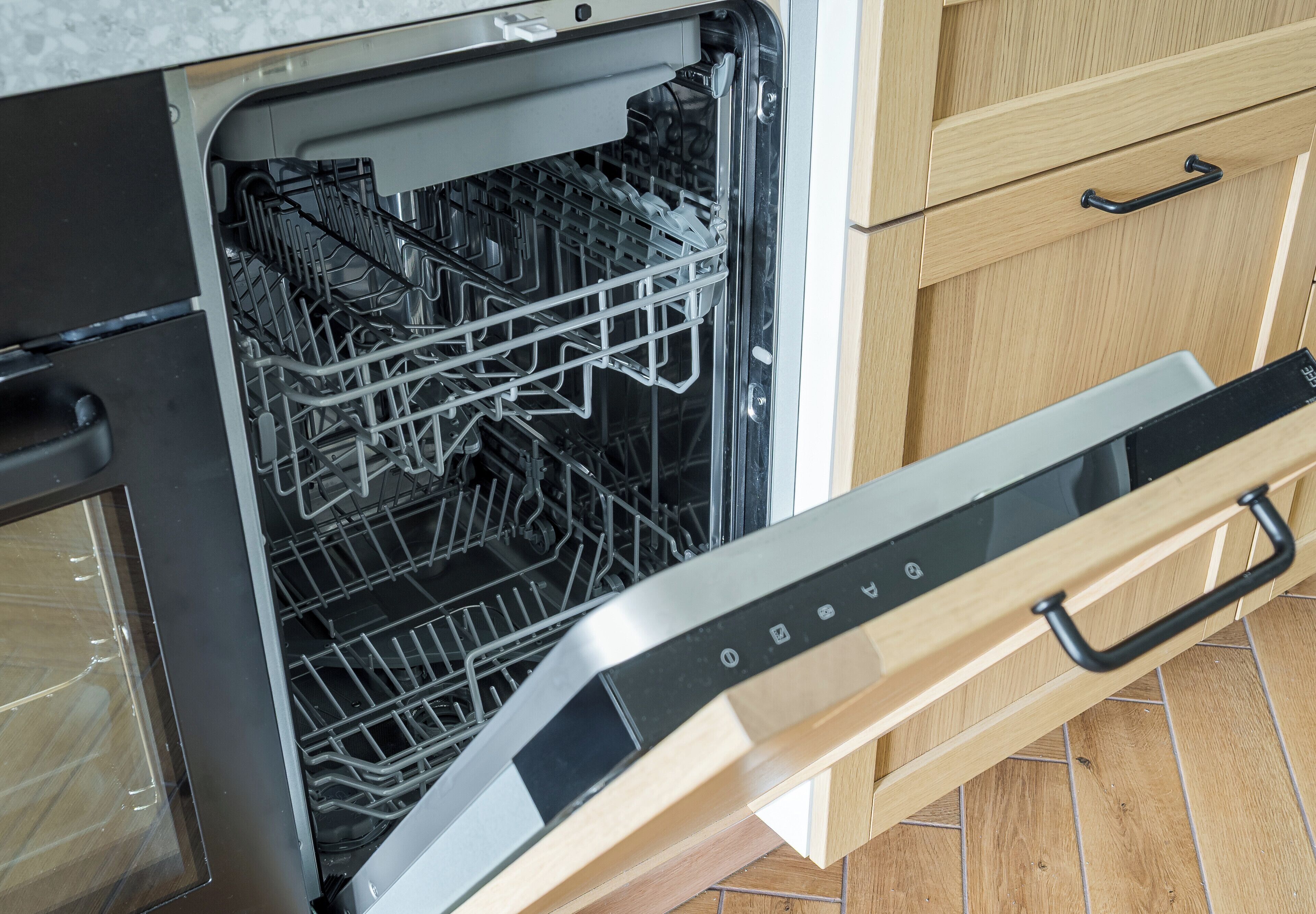 An open dishwasher with racks and a control panel, set in a kitchen with wooden cabinets and a tiled floor.