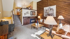 A modern living room with a fireplace, a staircase leading to an upper level, and a wooden wall with artwork. - Cedars 3: Ski-In/Out Peak 9 Townhome, Fireplace, Pool & Hot Tub Access. (Breckenridge)