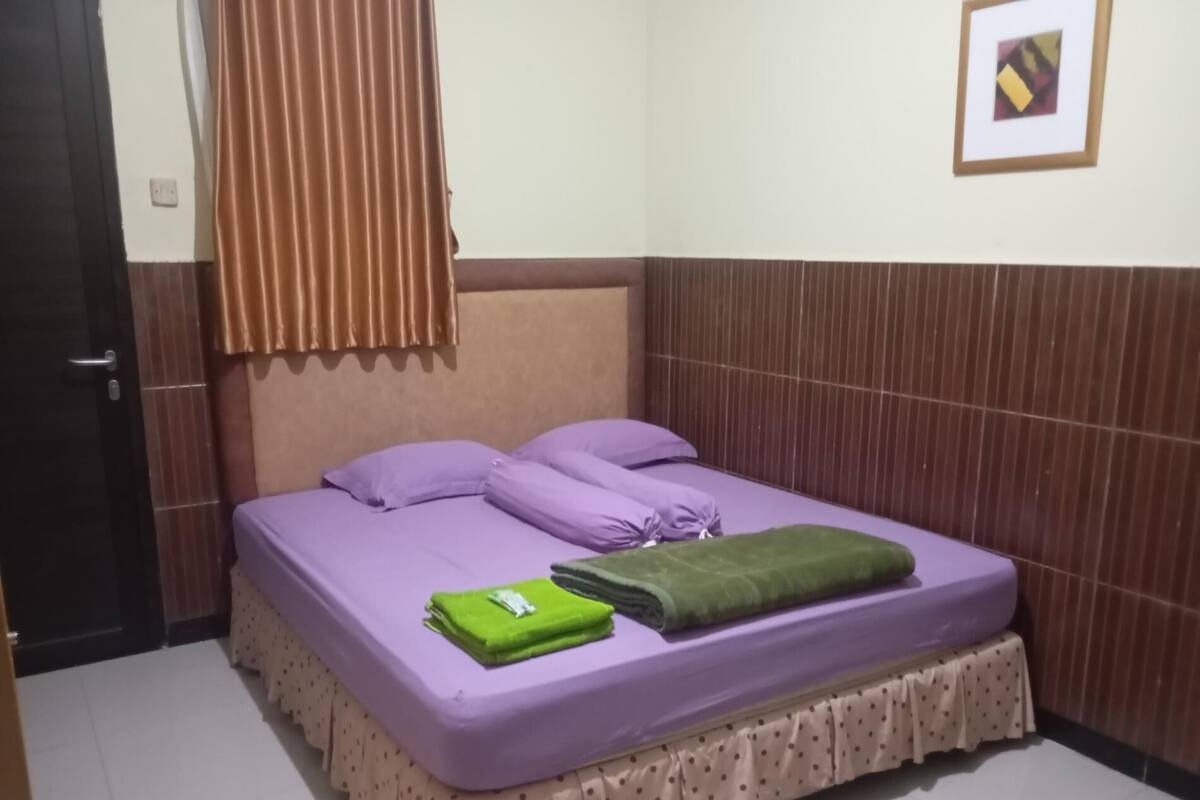 Una cama bien hecha con ropa de cama morada, una toalla verde y una cortina marrón.