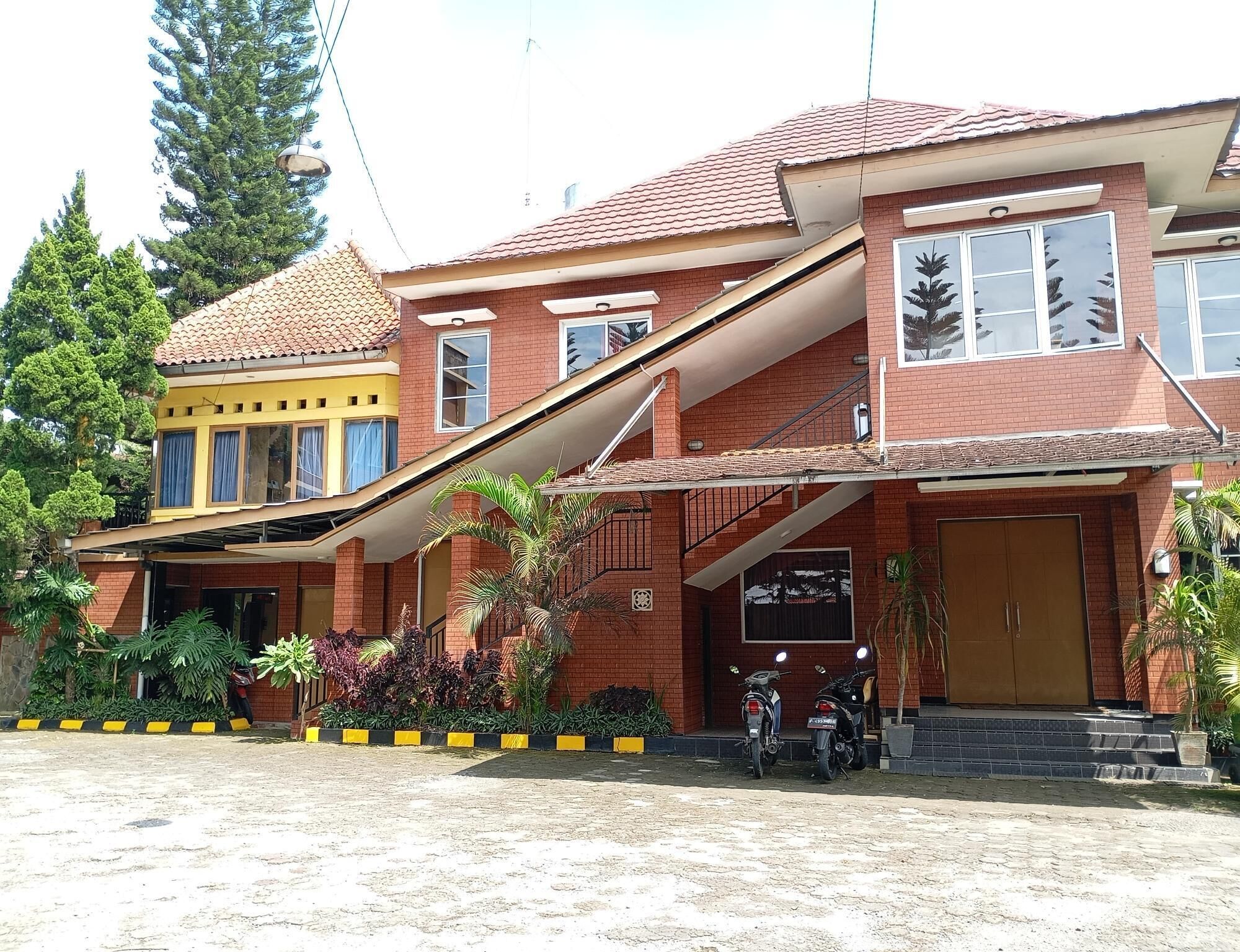 A two-story building with a red brick facade, a yellow section on the left, and a staircase leading to the upper floor. There are two motorcycles parked in front, and the entrance has a large wooden door.