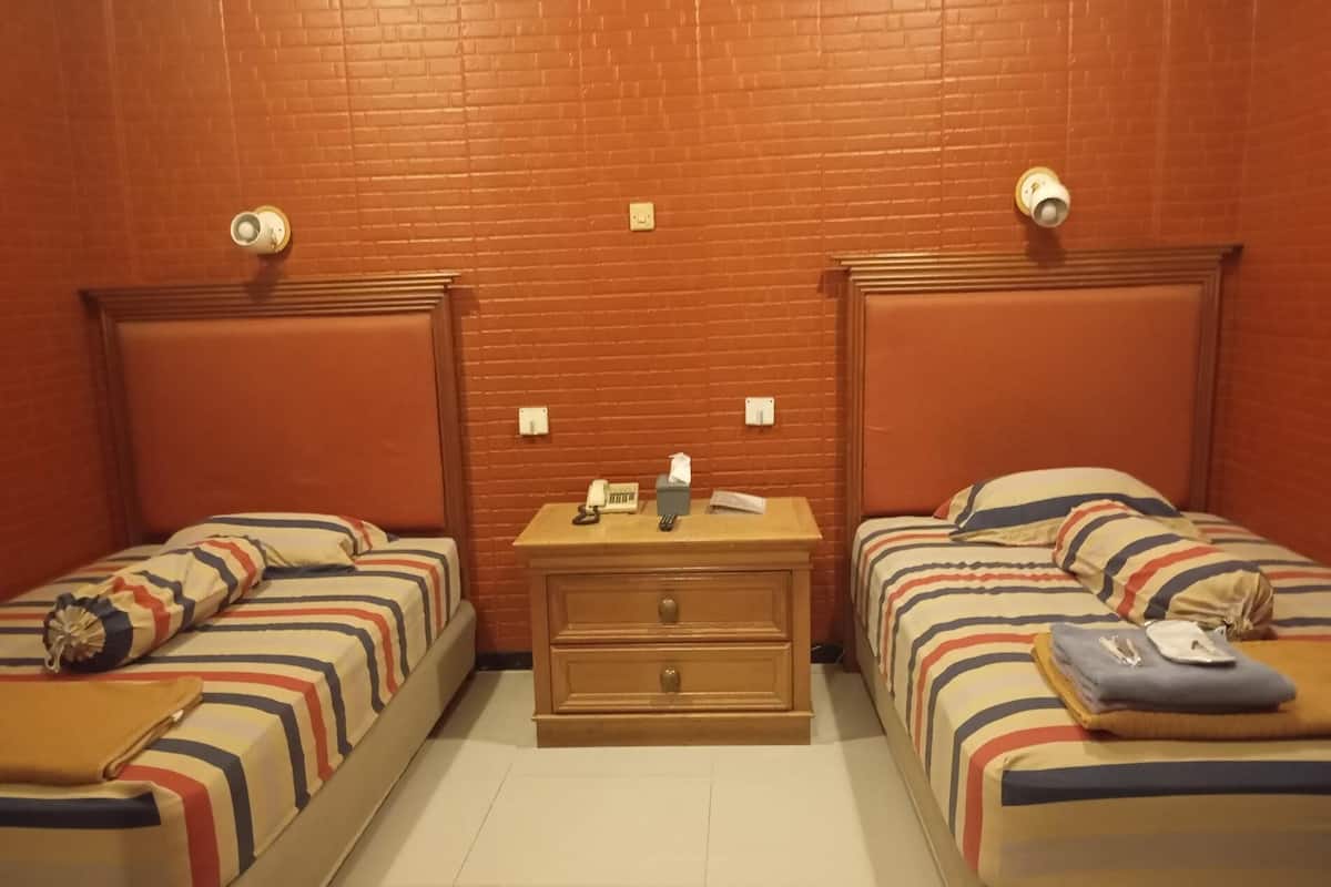 Una habitación con dos camas individuales, cada una con ropa de cama a rayas y una almohada. Entre las camas hay una mesita de noche de madera con una lámpara, una caja de pañuelos y un teléfono. Las paredes son de ladrillo, y hay dos interruptores de luz en la pared.