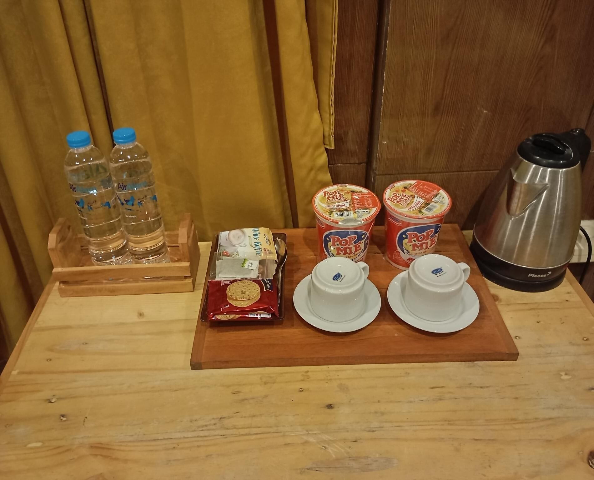 A wooden table with a tray containing water bottles, a pack of biscuits, two containers of PopMe tea, and a coffee maker.
