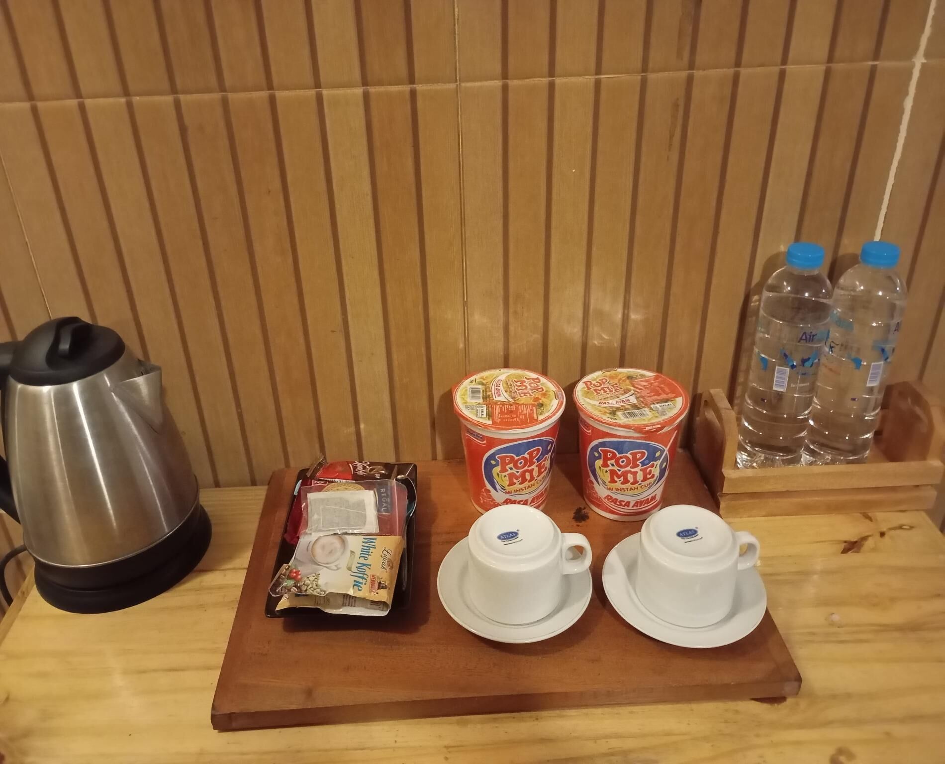 A wooden table with a kettle, instant noodles, a packet of white coffee, and two cups on a tray.