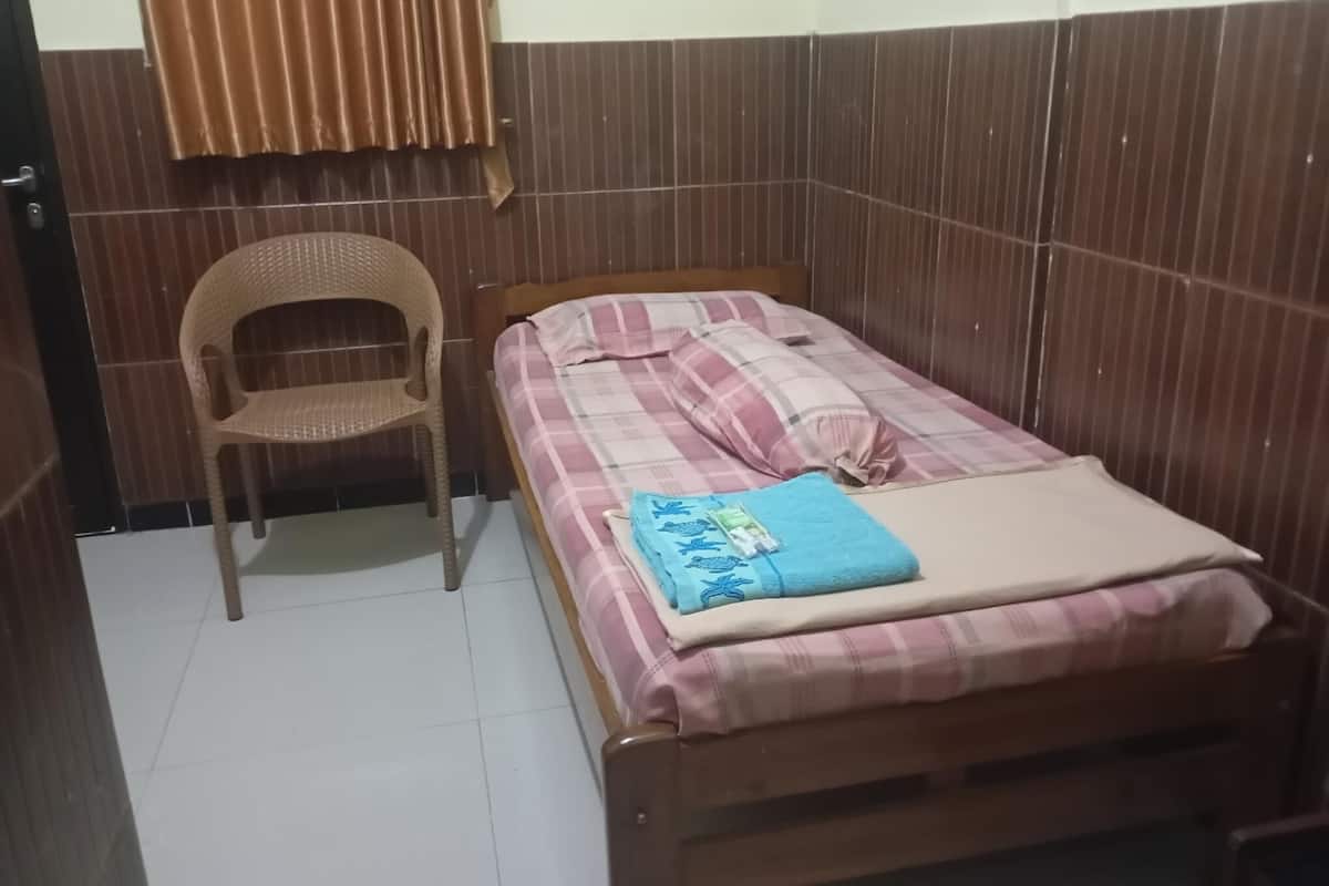 Una habitación pequeña con cama individual, una cama de madera, una silla de mimbre y una ventana con cortinas marrones.