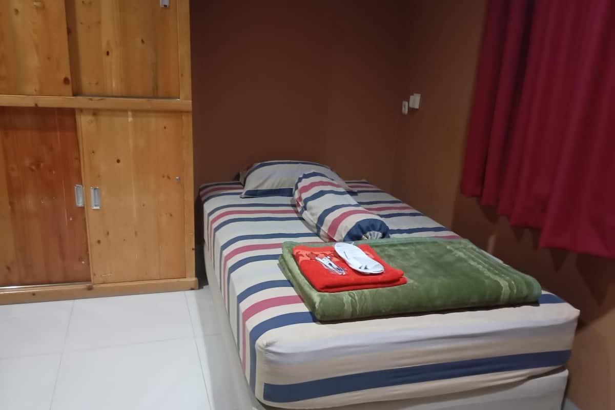 Una cama individual con ropa de cama a rayas, una almohada verde y una manta roja. Un armario de madera se encuentra a la izquierda de la cama.