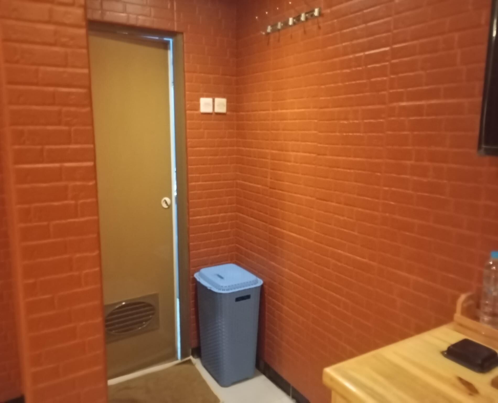 A narrow hallway with brick walls, a door at the end, a blue trash bin, and a wooden bench with a phone on it.
