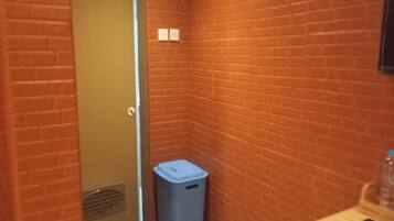 A narrow hallway with brick walls, a door at the end, a blue trash bin, and a wooden bench with a phone on it.