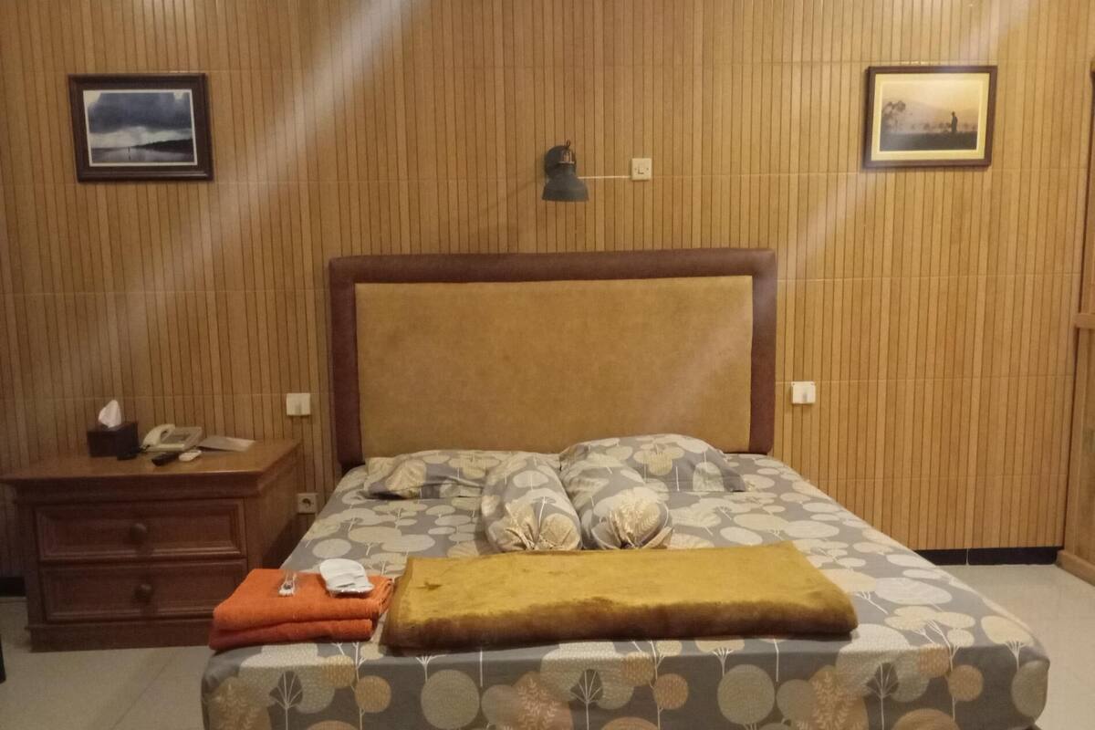 Un dormitorio con pared de paneles de madera, una cama con ropa de cama estampada, una mesita de noche con lámpara y dos cuadros enmarcados.