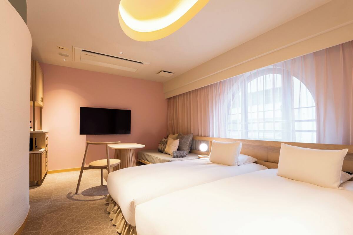 A hotel room with a large bed, a desk, a flat-screen TV, and a large window with sheer curtains.