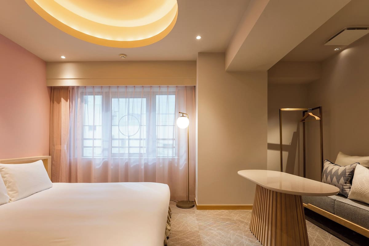 A modern hotel room with a large bed, a round table, a sofa, and a large window with sheer curtains.