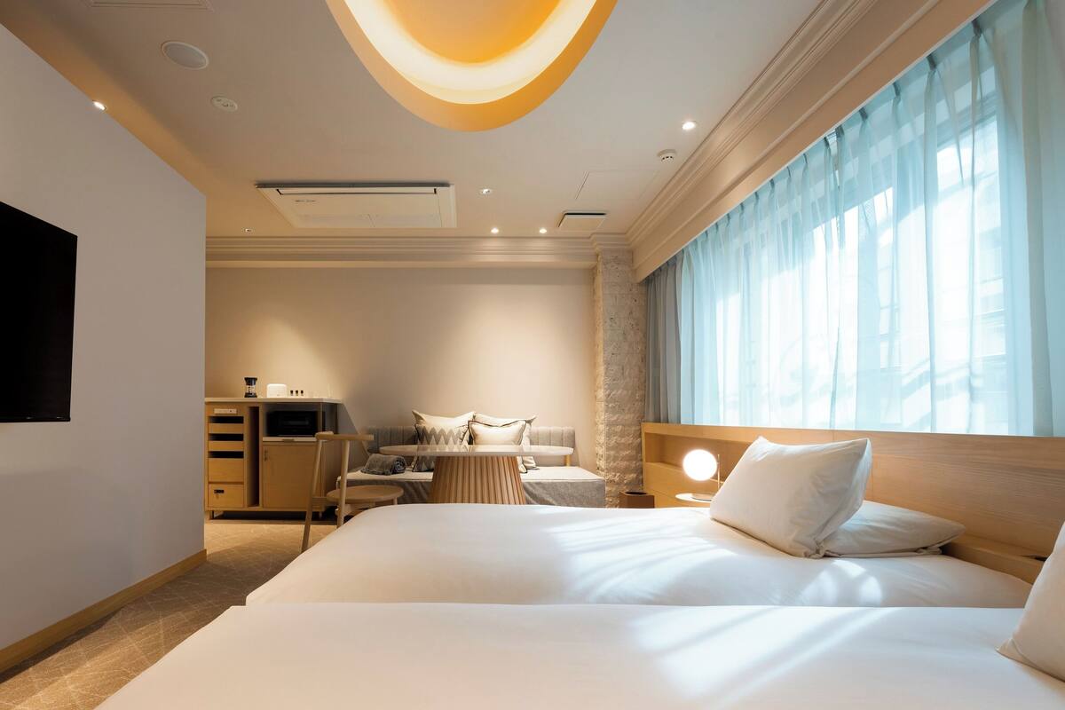 A hotel room with a large bed, a flat-screen TV mounted on the wall, a desk with a lamp, and a view of the city through large windows.