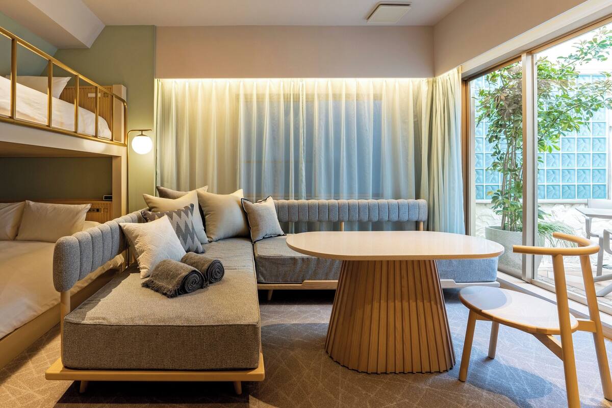 A modern hotel room with a bunk bed, a sofa, a round table, and a view of greenery outside.