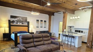 A modern living room with a brown leather sofa, a glass coffee table, and a colorful rug. There is a kitchen area with white cabinets and bar stools.