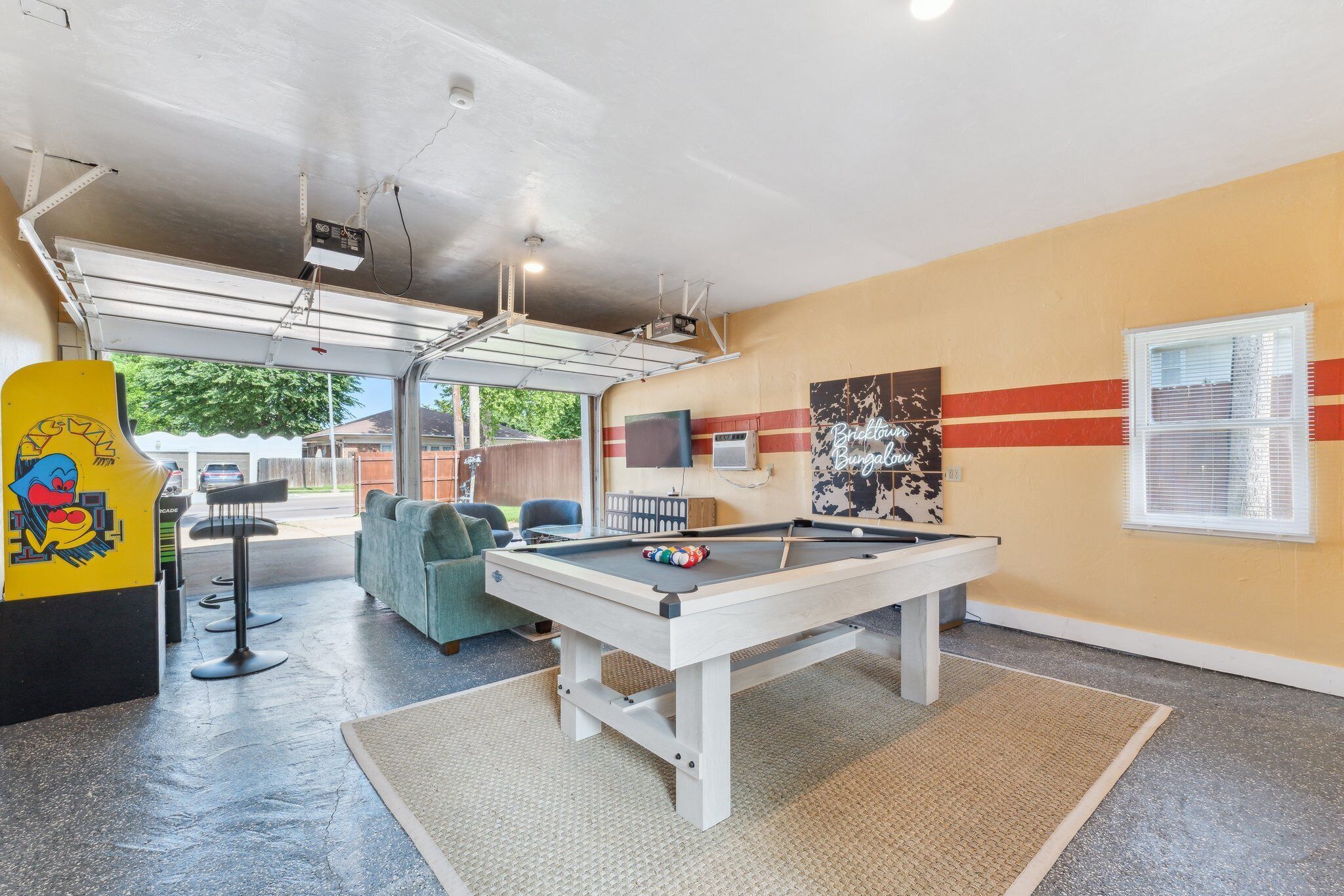 A pool table in a room with a yellow wall, a mounted TV, and a window with blinds.