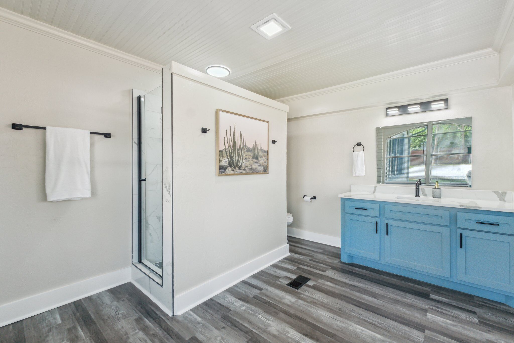 Un baño con cabina de ducha de vidrio, inodoro blanco, tocador azul con espejo y un cuadro en la pared.