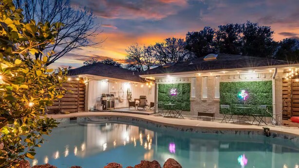 A swimming pool with a landscaped area, a covered patio with seating, and a neon sign. - Mini Golf | Pool | Game room | Stylish & Spacious | Wifi | Free Parking (Dallas)