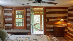 A rustic log cabin bedroom with a bed, a nightstand, a ceiling fan, and a window with a view of greenery outside.