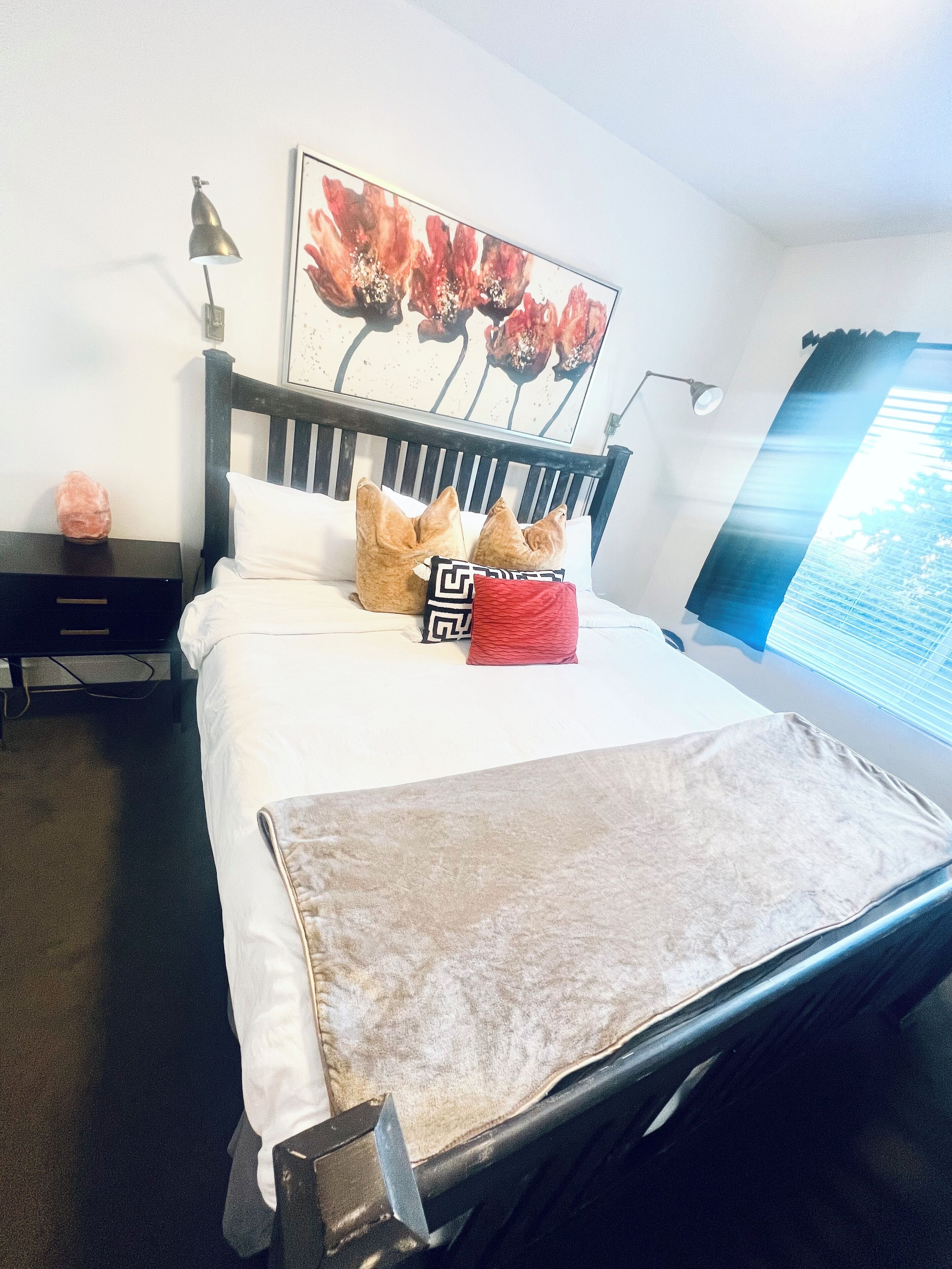 Uma cama bem arrumada com cabeceira, luminária de cabeceira, travesseiro decorativo e um quadro de flores na parede.