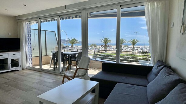 Een woonkamer met een bank, een televisie, een tafel en een balkon met uitzicht op zee en palmbomen.