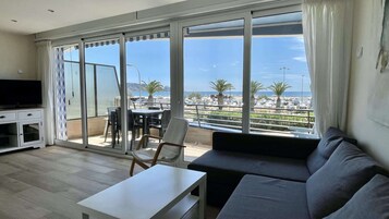 Una sala de estar con sofá, televisión, mesa y balcón con vistas al mar y palmeras.