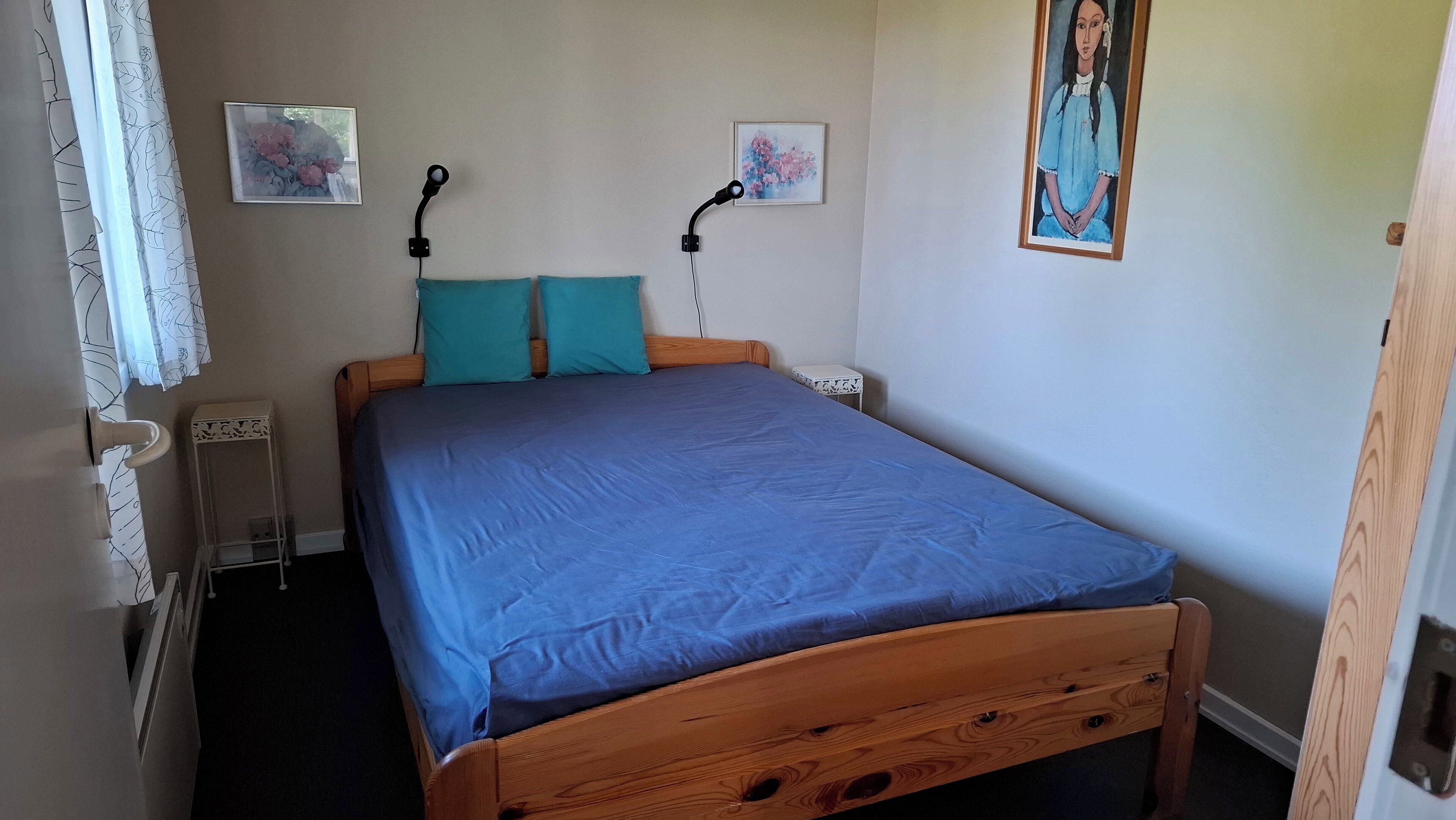 A bedroom with a wooden bed, two pillows, a nightstand, a wall-mounted lamp, and two framed artworks.