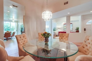 A modern living room with a glass-top coffee table, tufted chairs, a chandelier, and a view of the dining area.
