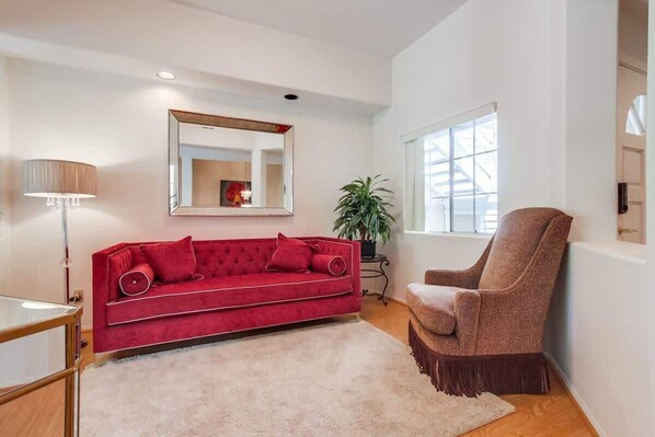 Una sala de estar con un sofá rojo, un sillón marrón, un espejo grande y una ventana con persianas.