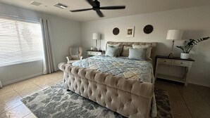 Une chambre à coucher avec un lit capitonné, un ventilateur de plafond, une table de chevet avec une lampe, une chaise et une fenêtre avec des stores.