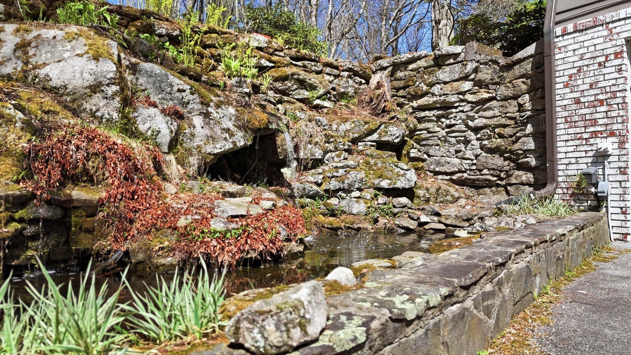 A stone retaining wall with a small stream, surrounded by vegetation and a brick building.