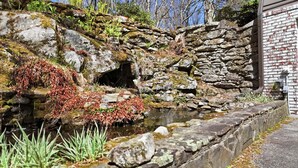 A stone retaining wall with a small stream, surrounded by vegetation and a brick building. - The Bodhi: Spacious 5-bedroom retreat in Highlands with wooded mountain views. (Highlands)