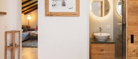 A modern bathroom with a wooden vanity, a large mirror with a circular light, and a framed picture on the wall.