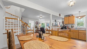 A spacious dining area with a wooden table and chairs, a kitchen with wooden cabinets and a window, and a staircase with white railings.