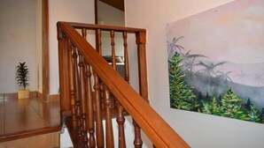 Una escalera de madera con barandilla, una planta en maceta y un cuadro con motivos tropicales en la pared.
