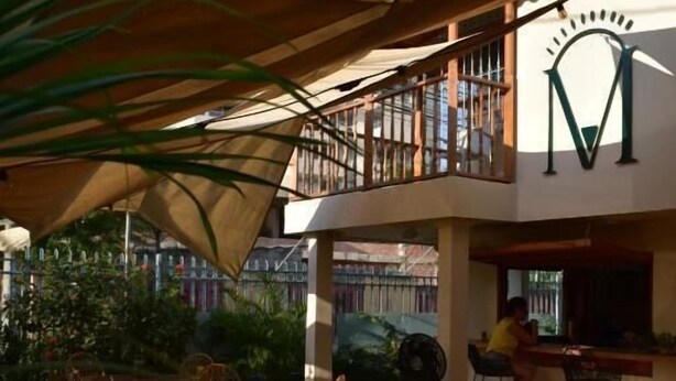 Un área de estar al aire libre con mesas y sillas, un balcón cubierto y un gran letrero con la letra 'M'.
