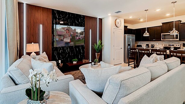 Ein modernes Wohnzimmer mit einer Sofagarnitur, einem Couchtisch und einem an der Wand montierten Fernseher. Es gibt eine Küchenzeile mit einer Mikrowelle und einem Kühlschrank.