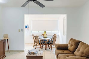 A living room with a brown sofa, a dining table with chairs, a ceiling fan, and a window with blinds.
