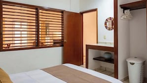 Un dormitorio con cama, una puerta de madera, una ventana con persianas, una mesita con un florero y un cubo de basura.