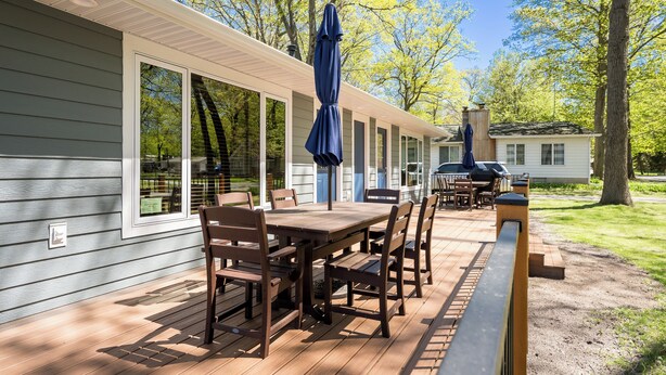 A wooden deck with a table and chairs, a covered umbrella, and a view of a house and trees. - Awesome Silver Lake Rental - Short Distance to Lake MI (Mears)