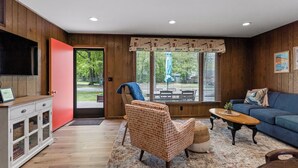 A living room with a wooden floor, a blue sofa, a patterned armchair, a coffee table, and a television on a cabinet. - Awesome Silver Lake Rental - Short Distance to Lake MI (Mears)