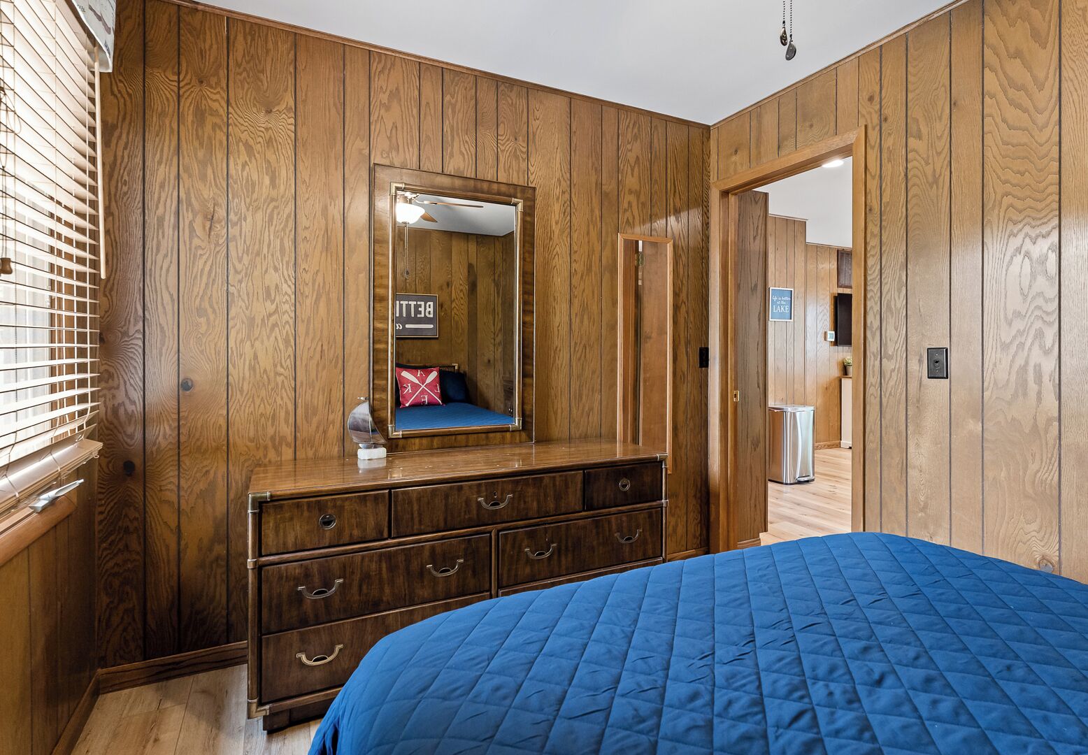Une chambre avec des lambris en bois, un grand lit recouvert d’une couette bleue, une commode avec des tiroirs, un miroir et un panneau indiquant la présence d’une salle de bain avec vue sur le lac.