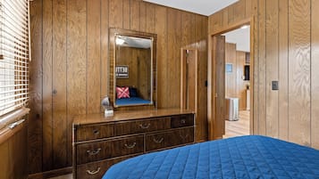 Une chambre avec des lambris en bois, un grand lit recouvert d’une couette bleue, une commode avec des tiroirs, un miroir et un panneau indiquant la présence d’une salle de bain avec vue sur le lac.