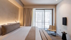 Een moderne hotelkamer met een groot bed, een stoel, een klein tafeltje en uitzicht door een groot raam.