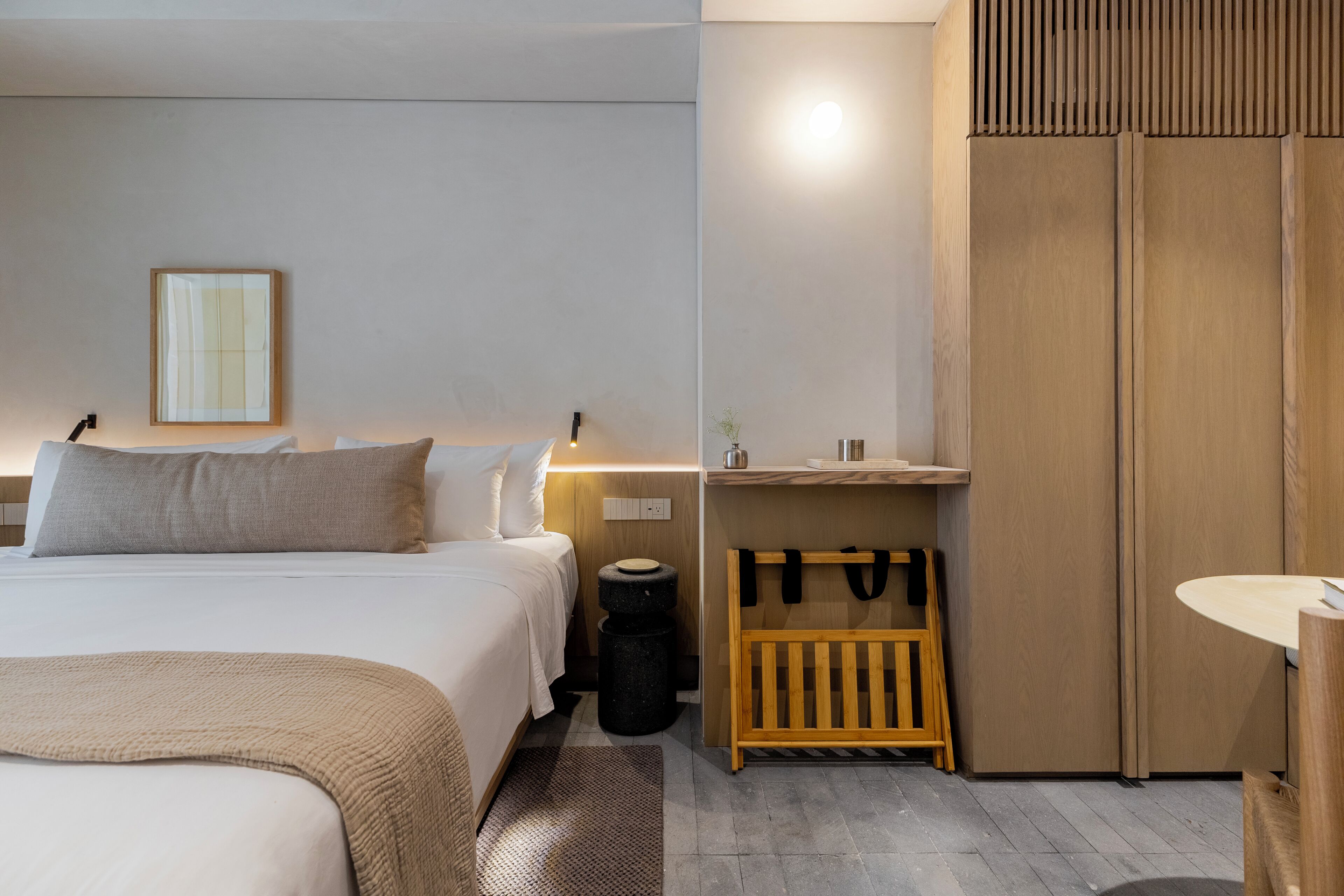 A modern hotel room with a bed, bedside table, lamp, and a wooden wardrobe.