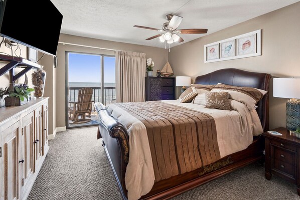 A bedroom with a large bed, a ceiling fan, a view of the ocean, and a balcony with a chair.